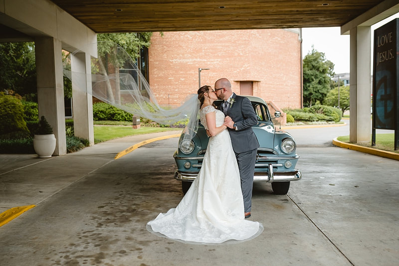 Bride and Groom kissing in front of vintage Plymouth with veil floating in the wind