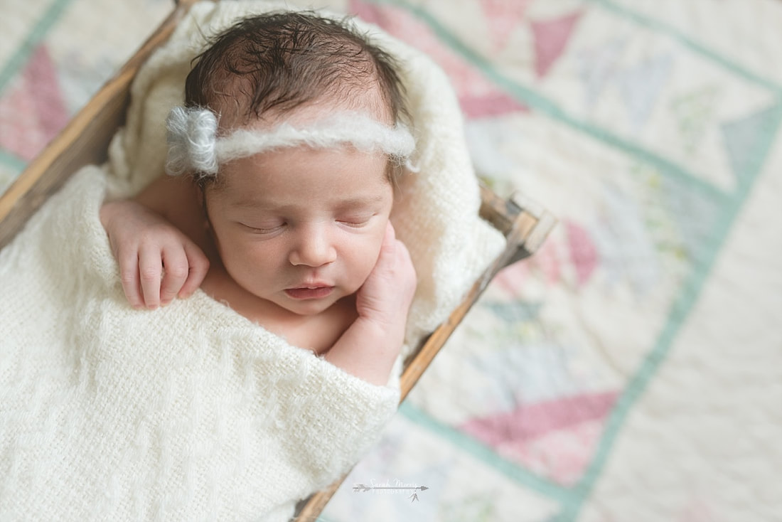 newborn baby girl wearing a bow, wrapped in a cream blanket, sleeping in a rustic wooden box on top of a vintage quilt