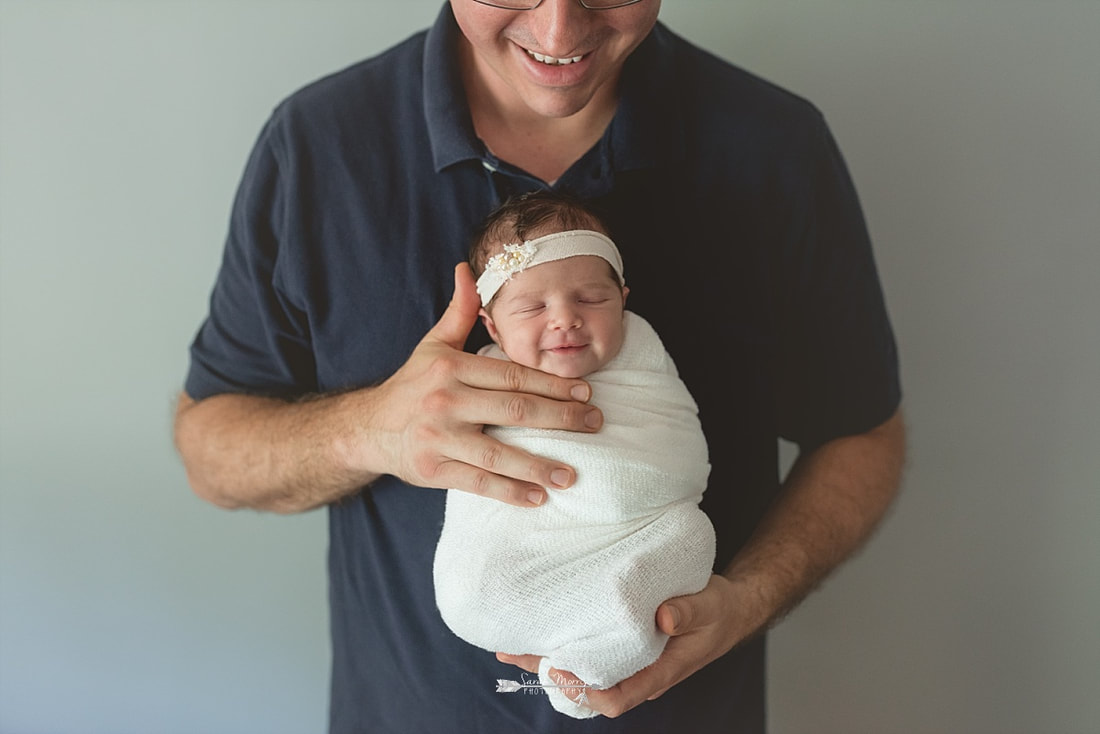 Newborn baby girl smiling in her daddy's hands and swaddled in a cream blanket