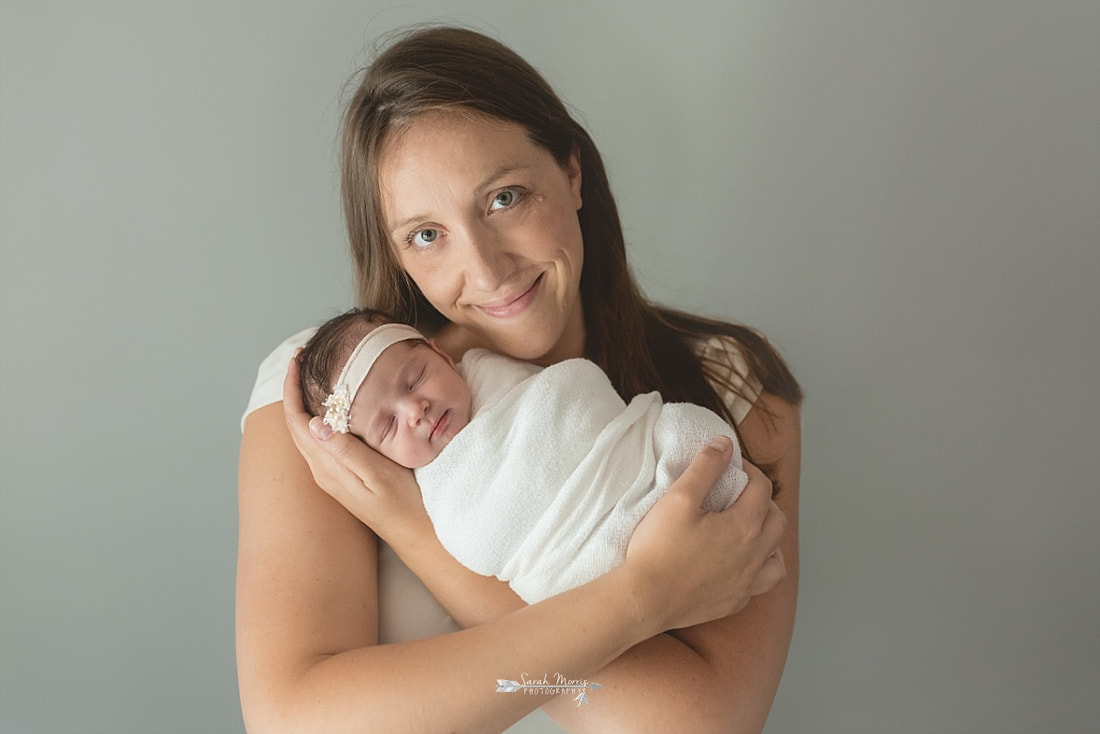 Mother holding her newborn baby girl close to her face while she's swaddled in a cream blanket