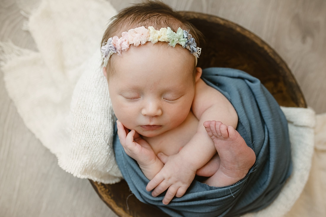 Baby girl wearing a rainbow headband swaddled in blue, sleeping in wooden bowl for newborn photoshoot in Memphis, TN